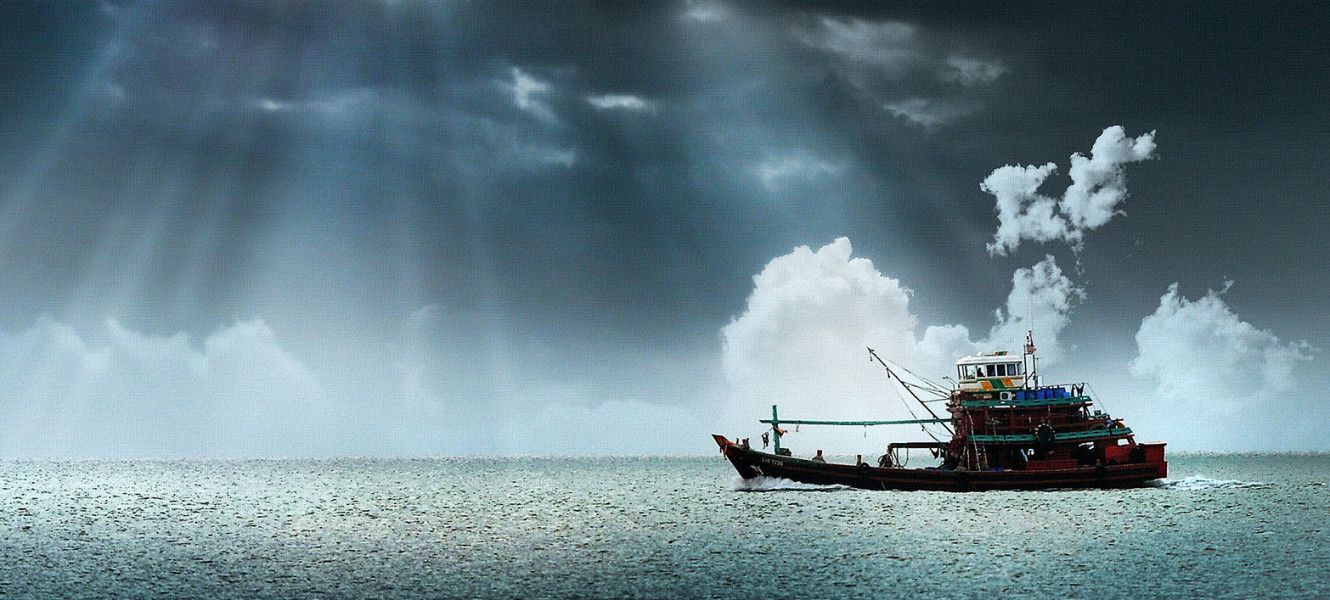 Fishing vessel at sea under a dark sky pierced with slanting sunbeams